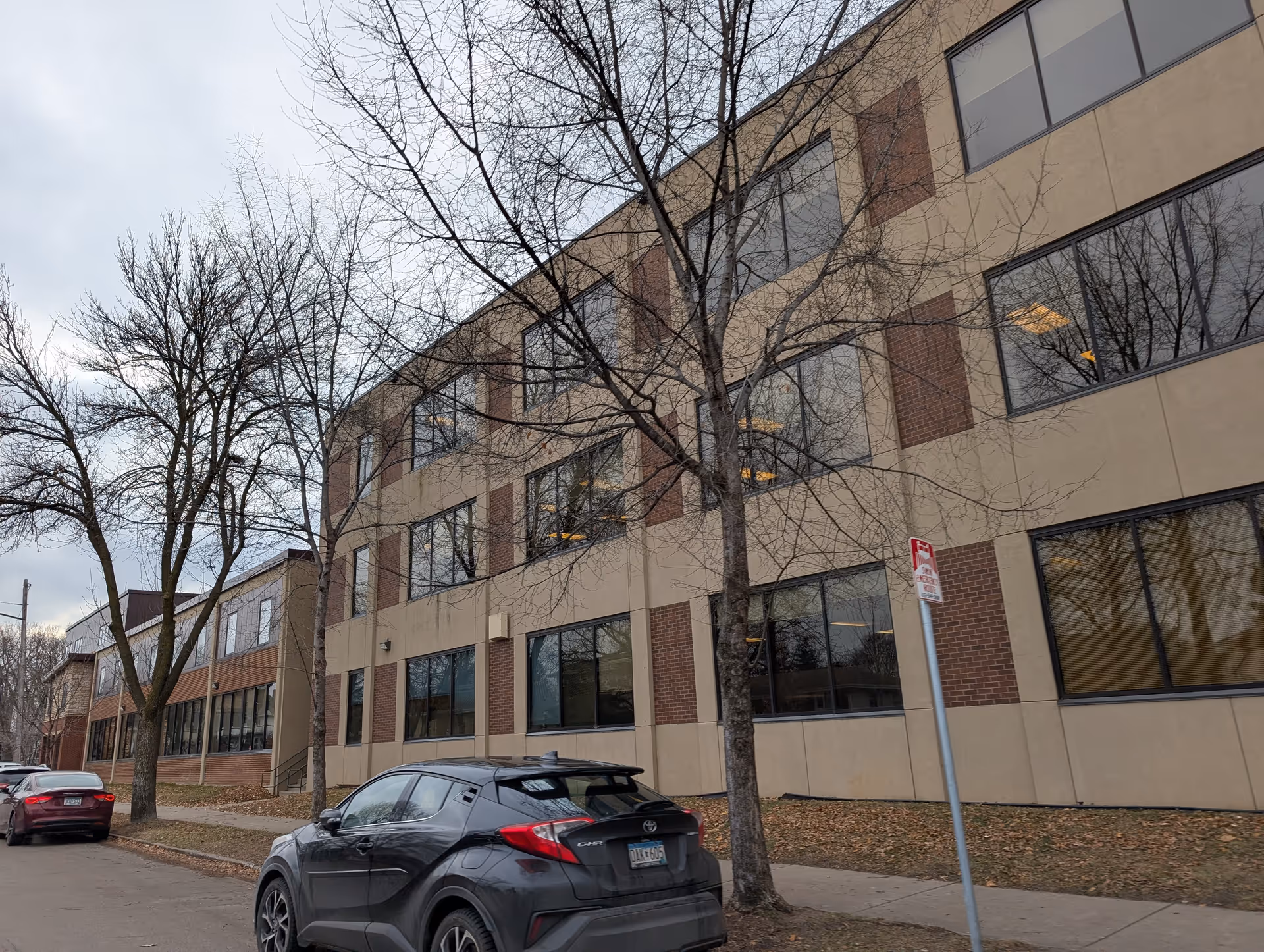 Exterior view of a three-story building with large windows and a beige and brick facade. Leafless trees line the sidewalk in front of the building, and several cars are parked along the street. The sky is overcast.