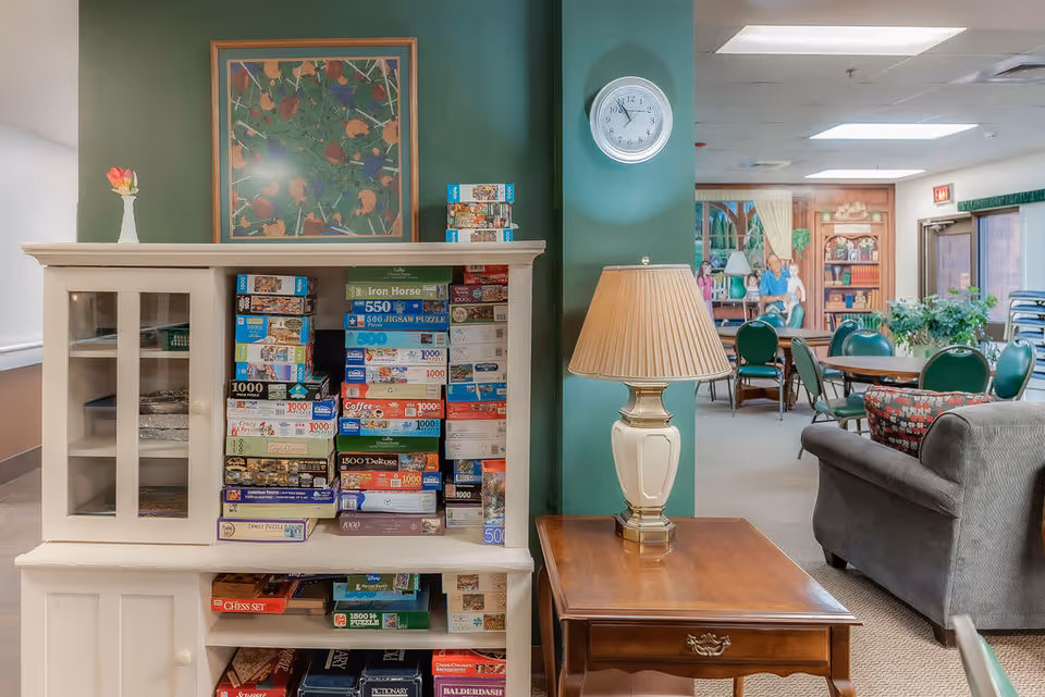 Interior view of a senior living facility common area with a white cabinet filled with various board games and puzzles, a wooden side table with a lamp, green walls, a clock showing 3:15, and a seating area with green chairs and a gray couch. A mural of people sitting in a library setting is visible in the background.