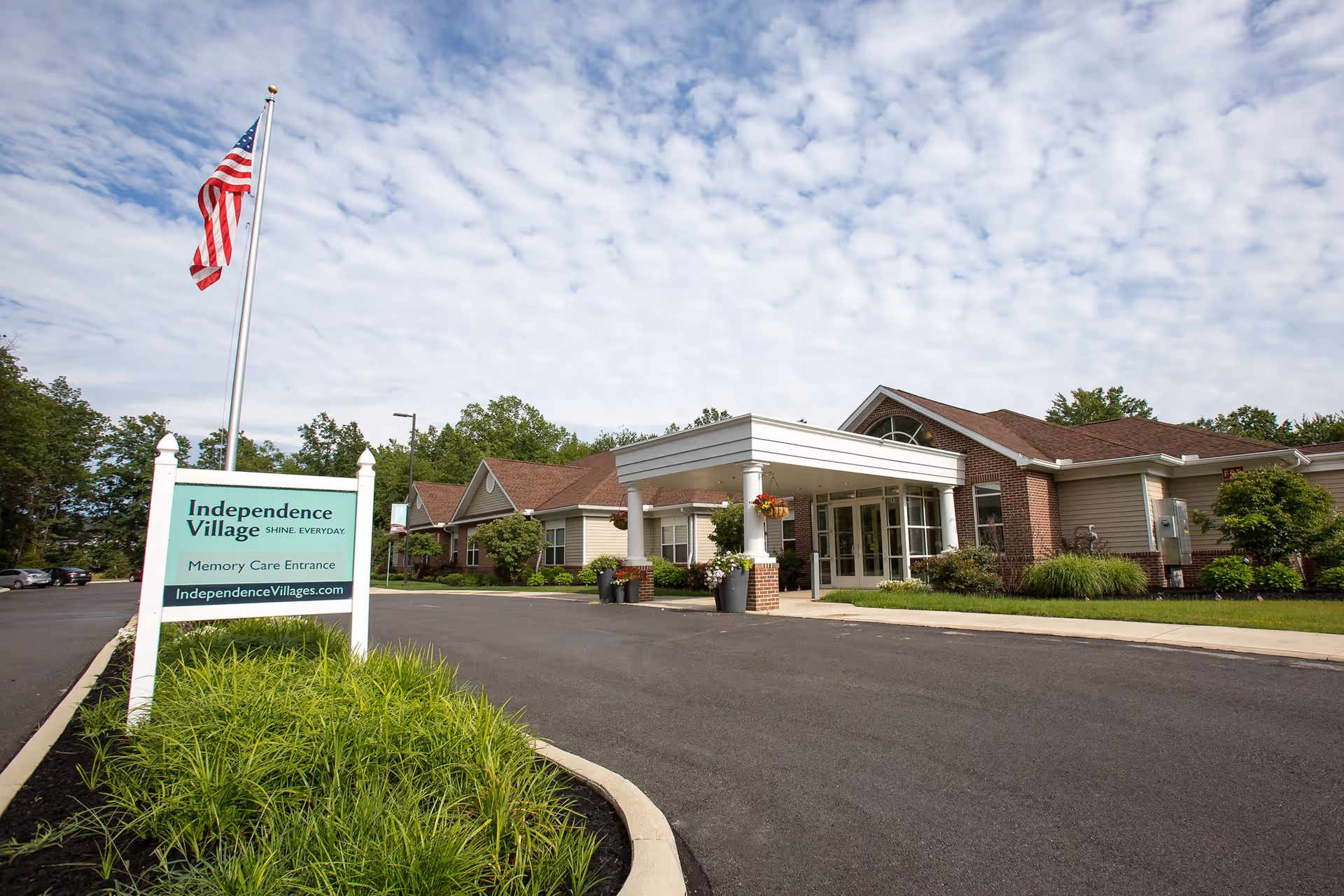 Exterior view of Independence Village of Avon Lake showing the entrance to the memory care section. The building has a covered entryway with white columns and hanging flower pots. There is a sign in the foreground with the facility's name and website, and an American flag flying on a flagpole. The sky is partly cloudy and there are trees and shrubs around the building.