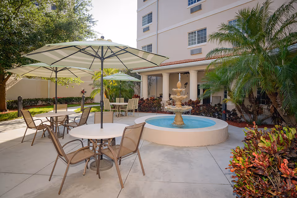 Outdoor patio area at Garden Plaza at Inverrary featuring round tables with umbrellas and chairs, a central tiered water fountain, surrounded by plants and trees, adjacent to a multi-story building.