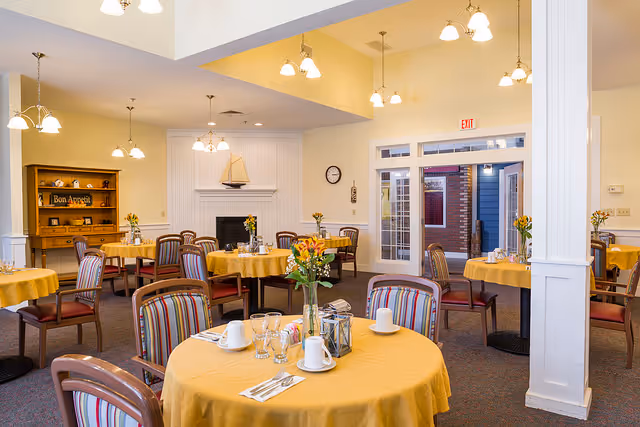 A bright dining room with round tables covered in yellow tablecloths, each set with cups, glasses, and silverware. The chairs have striped upholstery. The room features a white fireplace with a decorative sailboat on the mantel, a wooden hutch with a 'Bon Appetit' sign, and multiple hanging light fixtures. There is a door with glass panels leading outside and a clock on the wall.