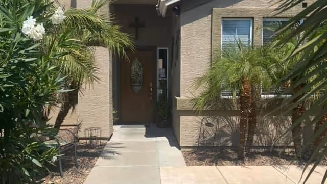 Concrete walkway leading to a stucco building's front door flanked by palm plants and a nearby window.