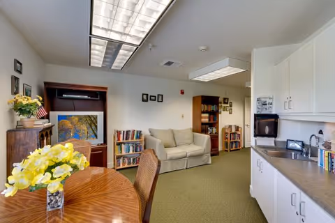 Well-lit senior living common area with a round table and flowers, kitchenette on the right, a sofa, bookshelves, and a TV cabinet.
