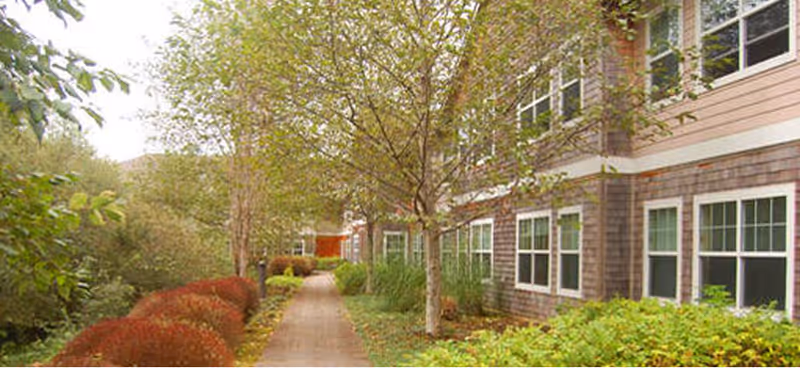 A paved walkway lined with trees and bushes runs alongside a two-story building with multiple windows. The building has a shingle exterior and is surrounded by greenery, including shrubs and small trees.