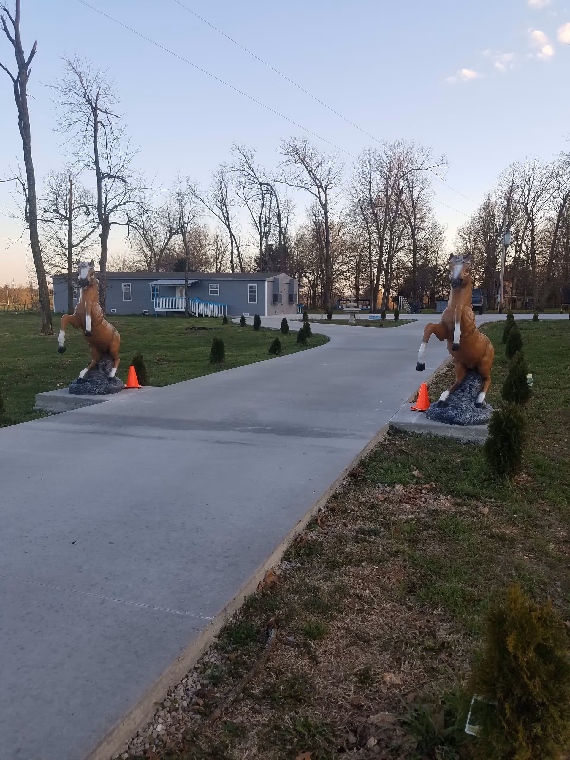 A paved driveway flanked by two rearing horse statues on concrete bases, each with an orange traffic cone nearby. The driveway leads to a single-story gray building with a small porch and ramp. The surrounding area has grass, small shrubs, and leafless trees under a clear sky.