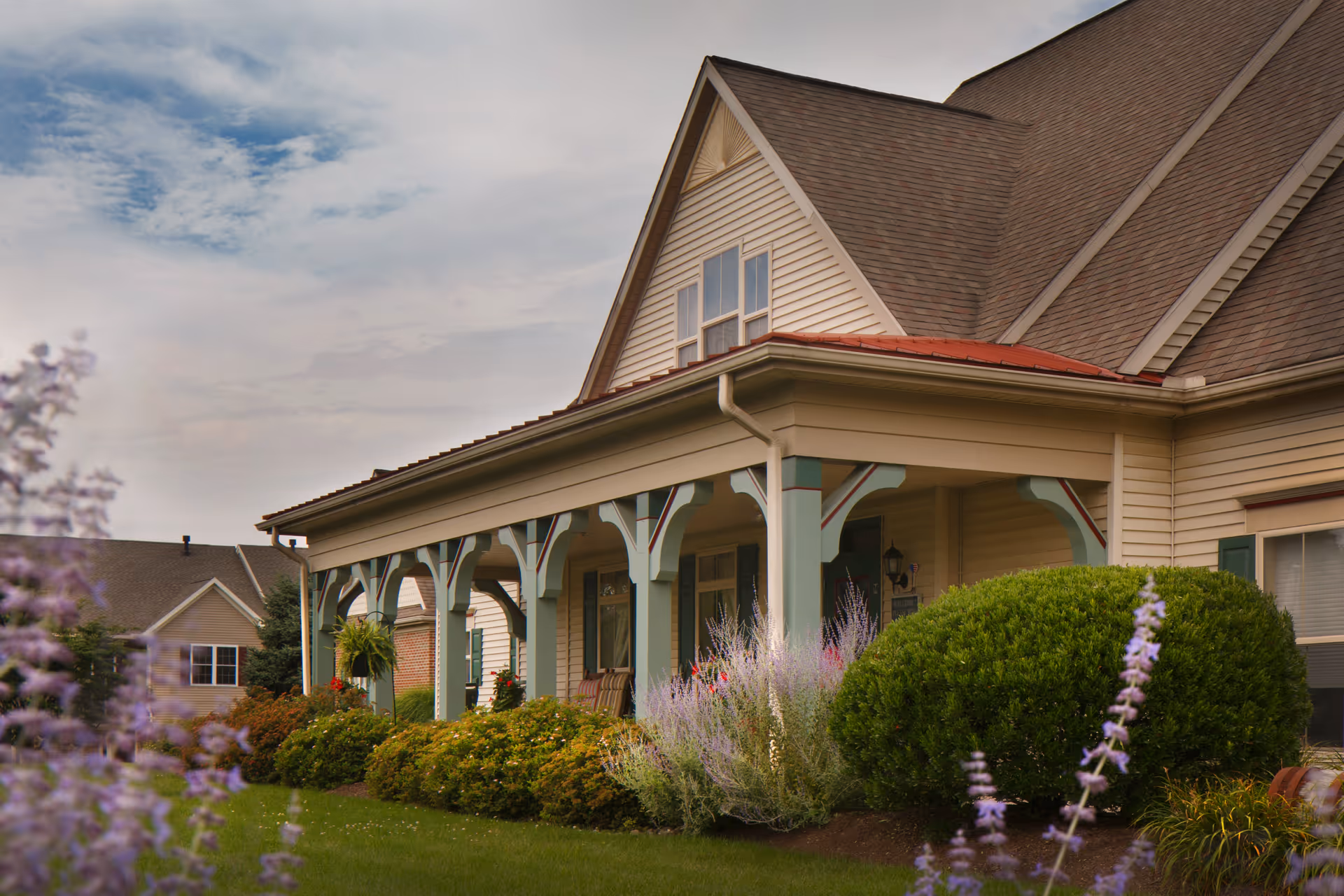 Exterior view of a residential-style building with a covered porch supported by decorative columns, surrounded by well-maintained bushes and flowering plants under a partly cloudy sky.