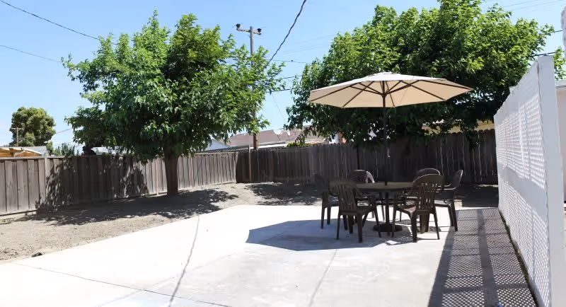 Outdoor patio area with a round table and four chairs under a large beige umbrella, surrounded by a wooden fence and trees providing shade.