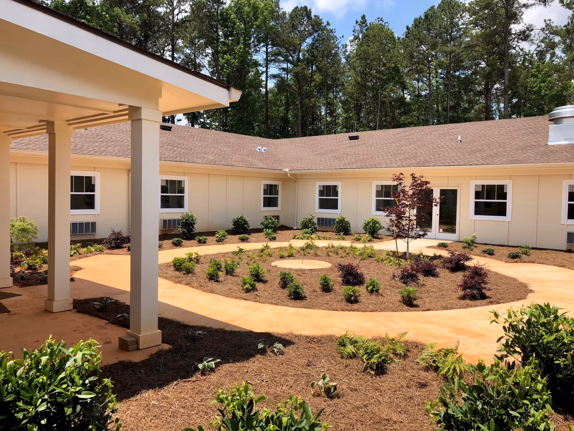 Outdoor courtyard area at Prema at Ashton Hills featuring a circular garden bed with small shrubs and a young tree in the center, surrounded by a paved walking path. The courtyard is enclosed by a single-story building with multiple windows and a glass door, with tall trees visible in the background under a partly cloudy sky.