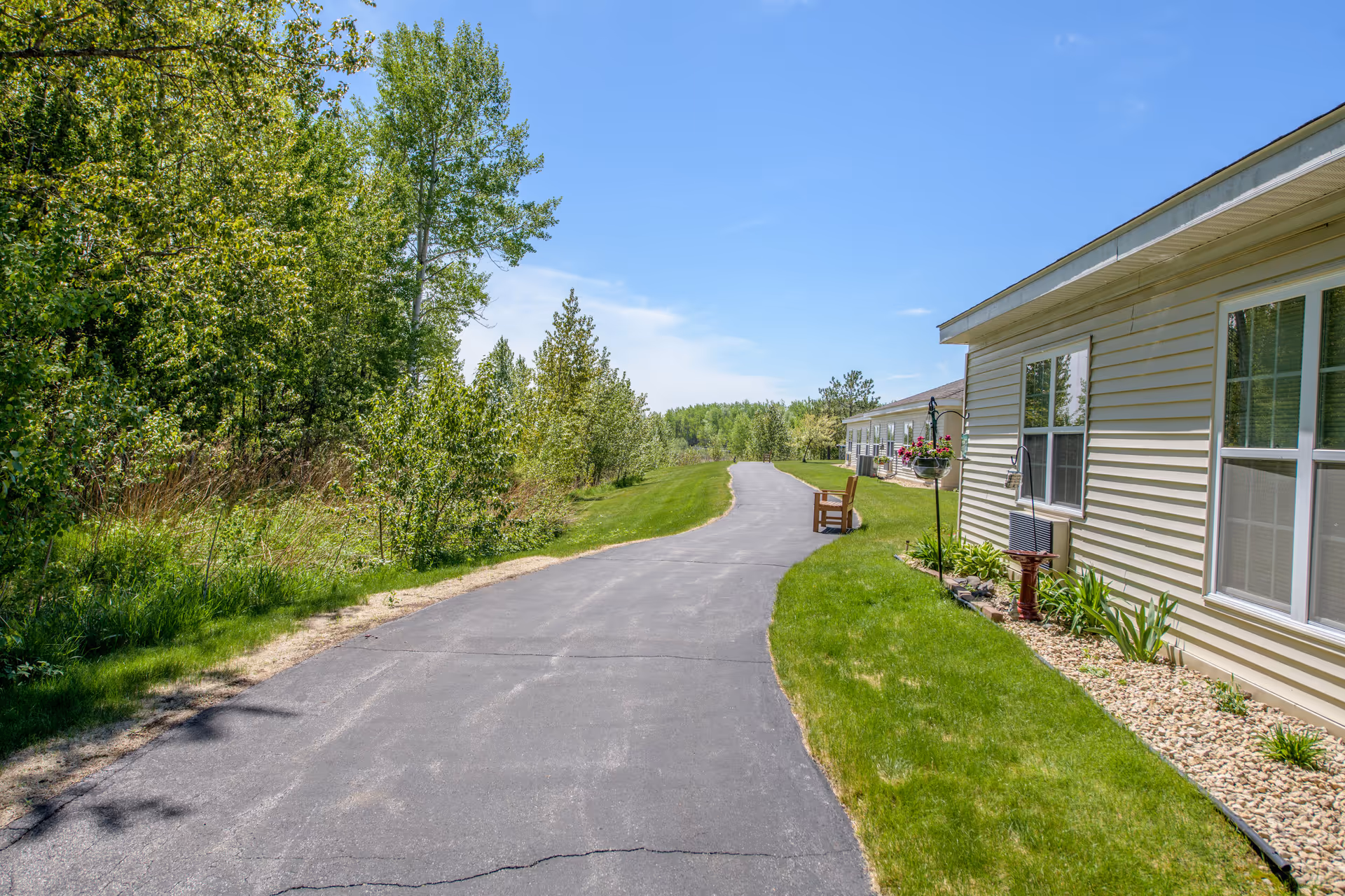 Paved walkway beside a light-colored building with lawn, landscaping, benches, and trees under a blue sky.
