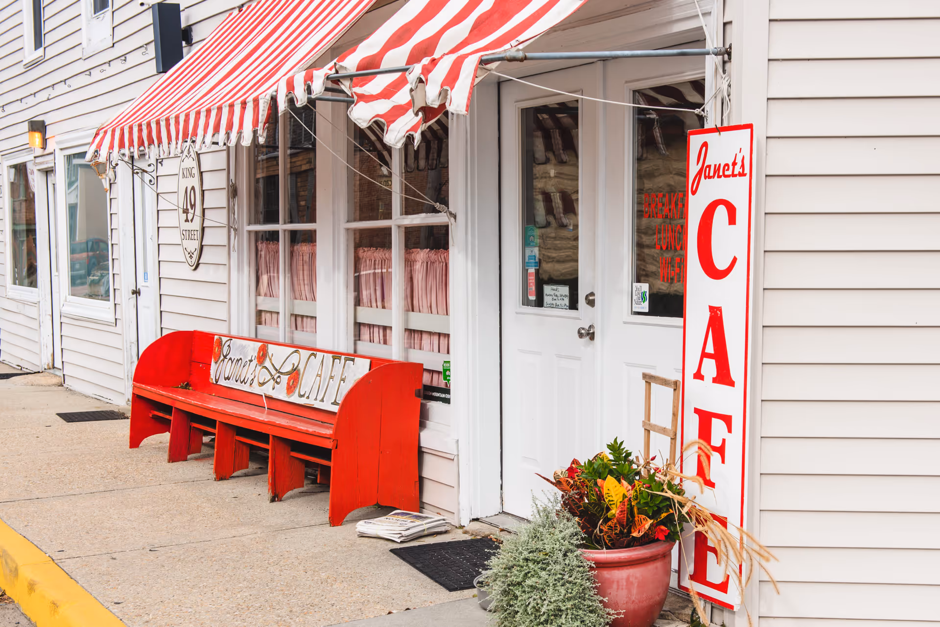 Exterior view of a cafe with a red and white striped awning, a red bench with a sign reading 'Janet's Cafe', a white door with glass panels, and a large vertical sign that says 'Janet's CAFE'. There are potted plants near the entrance and a sidewalk in front.