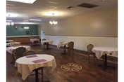 A dining room with several round tables covered with white tablecloths and purple placemats. The room has wooden flooring with a decorative circular inlay in the center. There are chairs around the tables and a buffet area with a countertop and cabinets along one wall. The walls are painted beige with white wainscoting, and ceiling lights provide illumination.