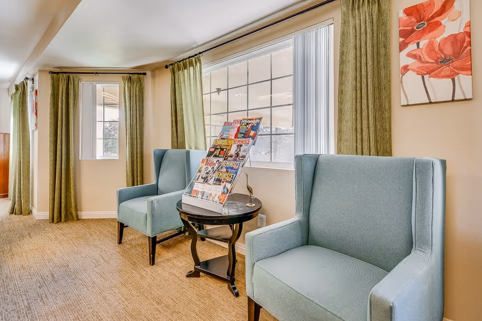 A cozy sitting area with two light blue armchairs separated by a small round black table holding a magazine rack filled with various magazines. The room has large windows with green curtains and beige walls adorned with a painting of red flowers.