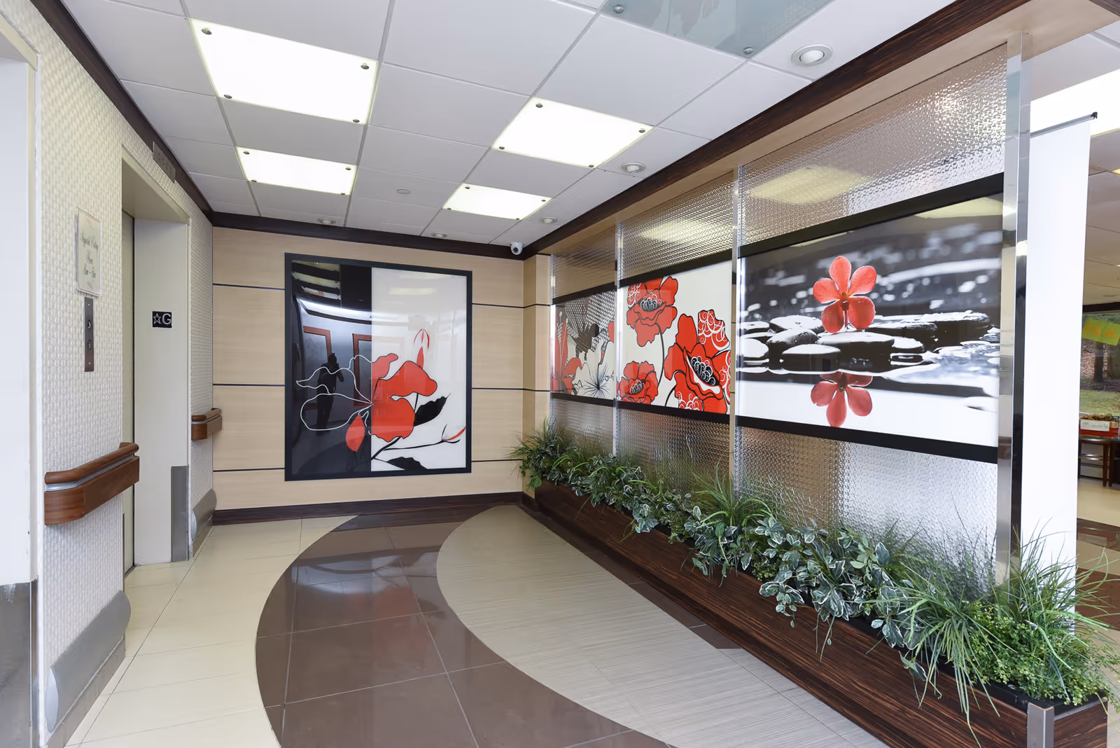 Interior hallway of a senior living facility with decorative wall art featuring red flowers and a planter with green plants along one side. The hallway has a tiled floor with a curved design and a ceiling with recessed lighting. There is an elevator door on the left side marked with a 'G' floor sign.