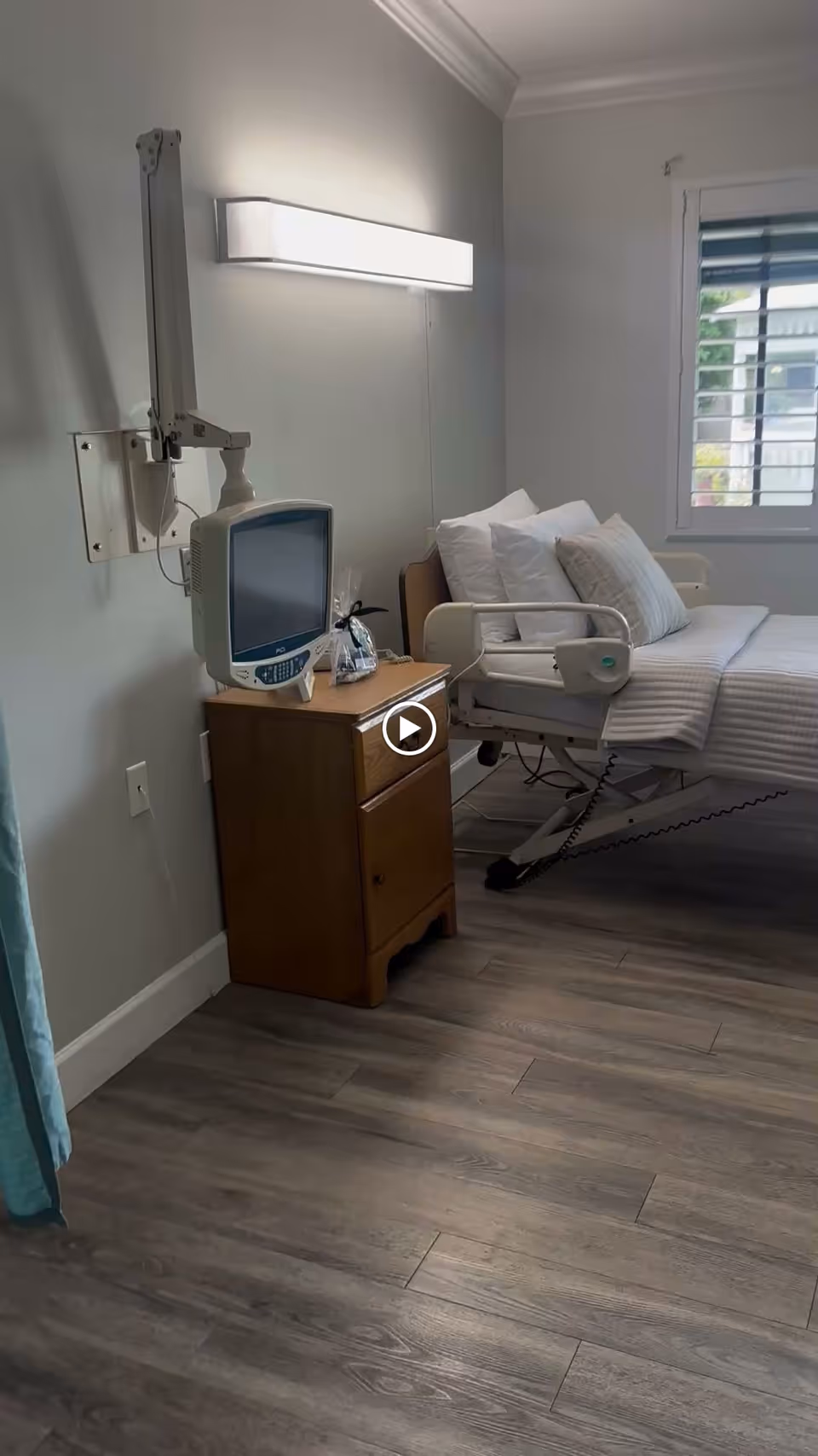 A tidy patient-style bedroom with a hospital bed, bedside cabinet holding a monitor, and a window with shutters.
