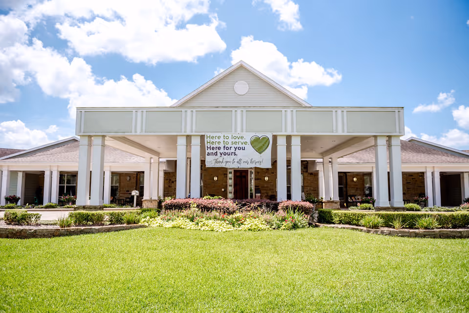 Front entrance of an assisted living facility with white columns, a covered porte-cochere, a banner above the doorway and a manicured green lawn.