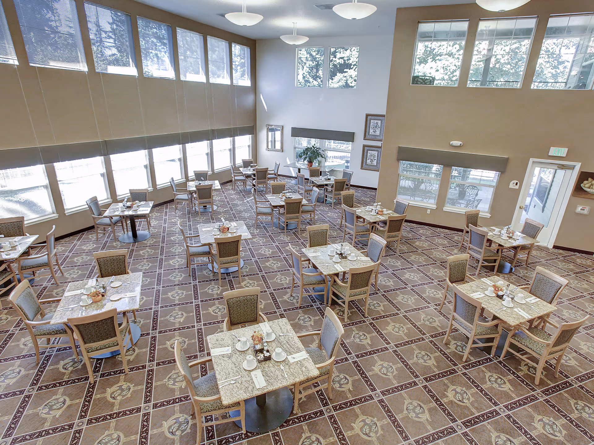A spacious dining room with multiple square tables each set with cups, plates, and utensils. The room has large windows with blinds, high ceilings, and patterned carpet flooring. The walls are painted in neutral tones and there are framed pictures and a potted plant near the windows.
