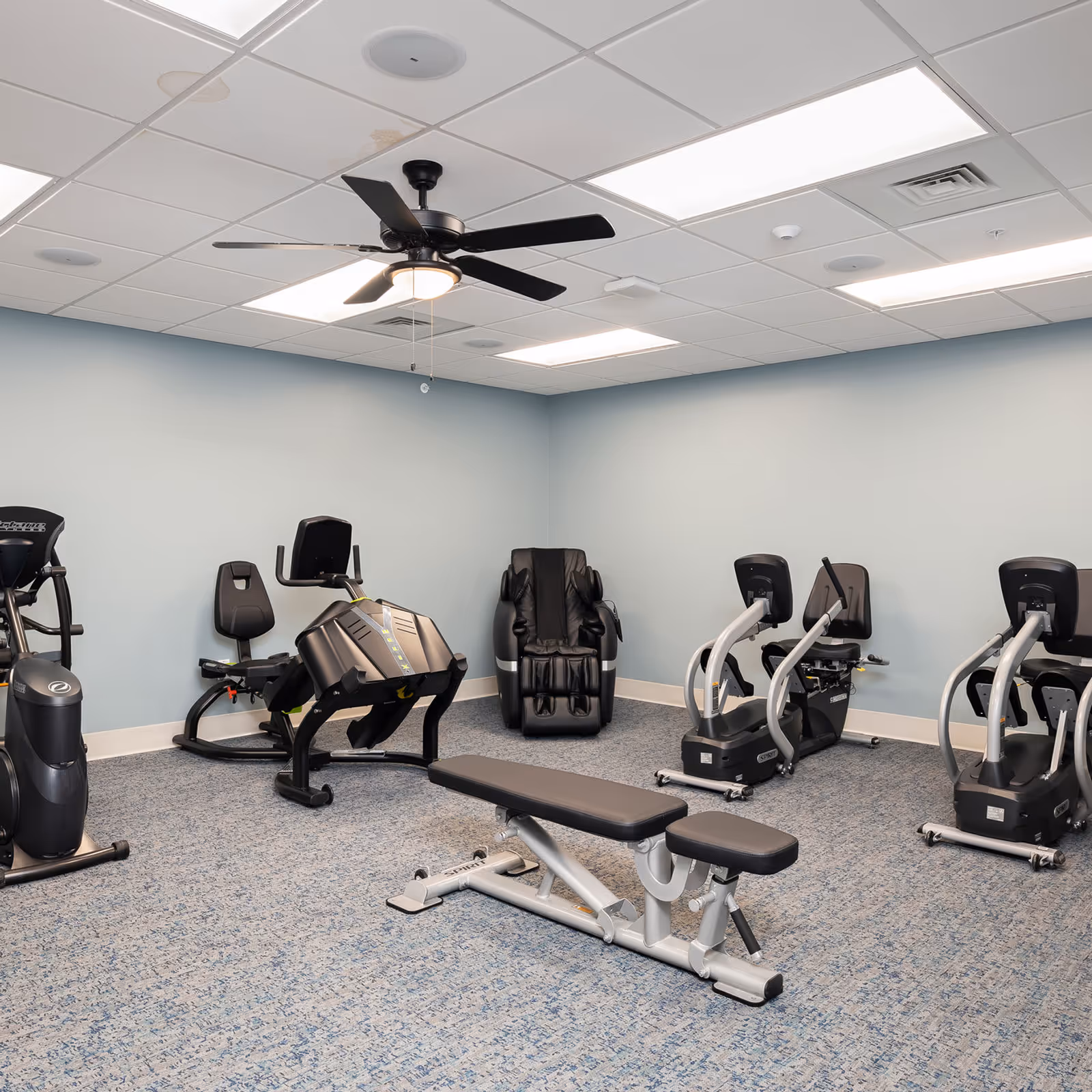 A senior living facility exercise room with various fitness equipment including stationary bikes, a leg press machine, a weight bench, and a black massage chair. The room has light blue walls, a patterned carpet, a ceiling fan, and fluorescent lighting.