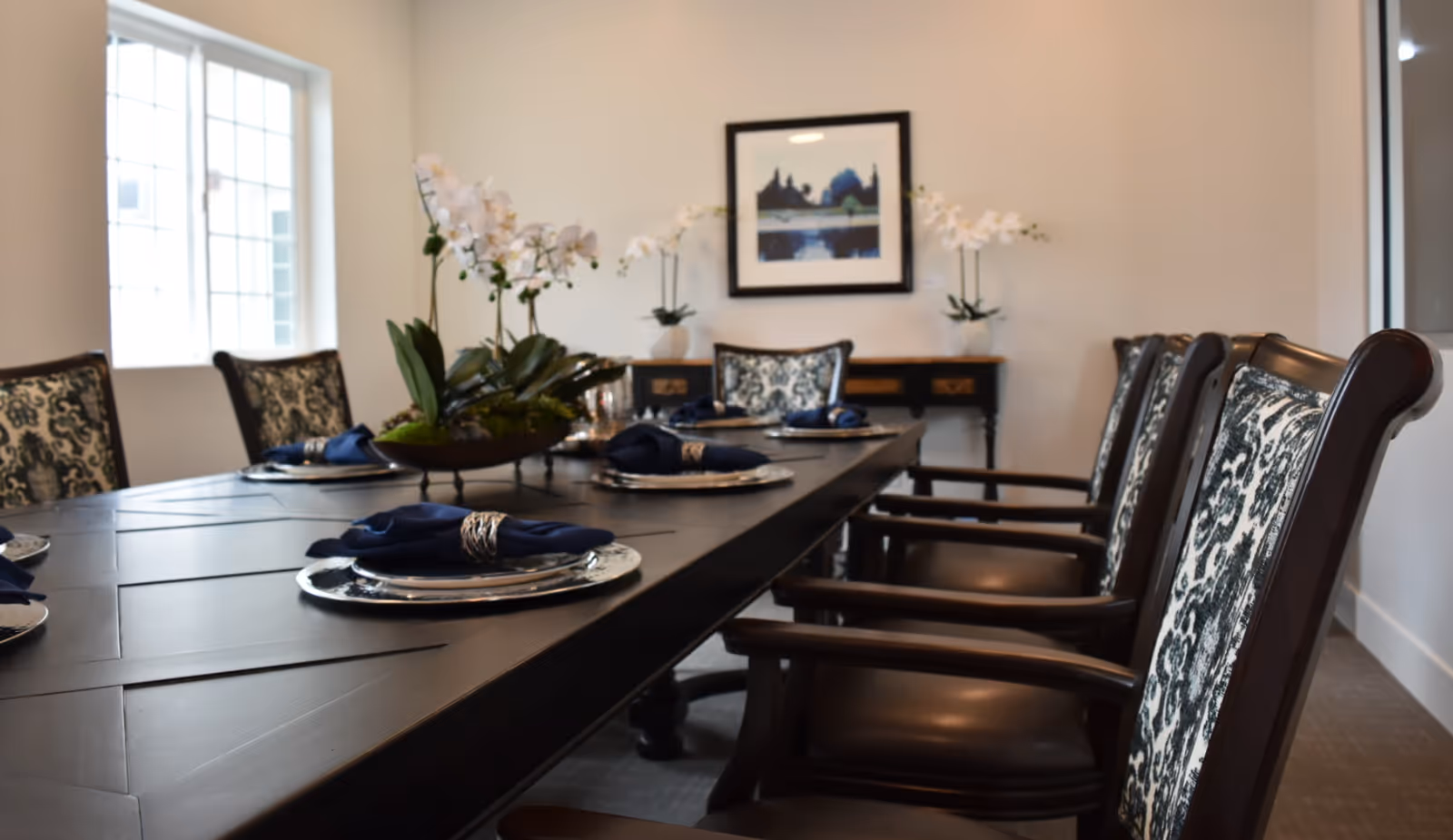 A formal dining room with a long dark table set with silver chargers and navy napkins, patterned chairs, orchids and framed art on the wall.