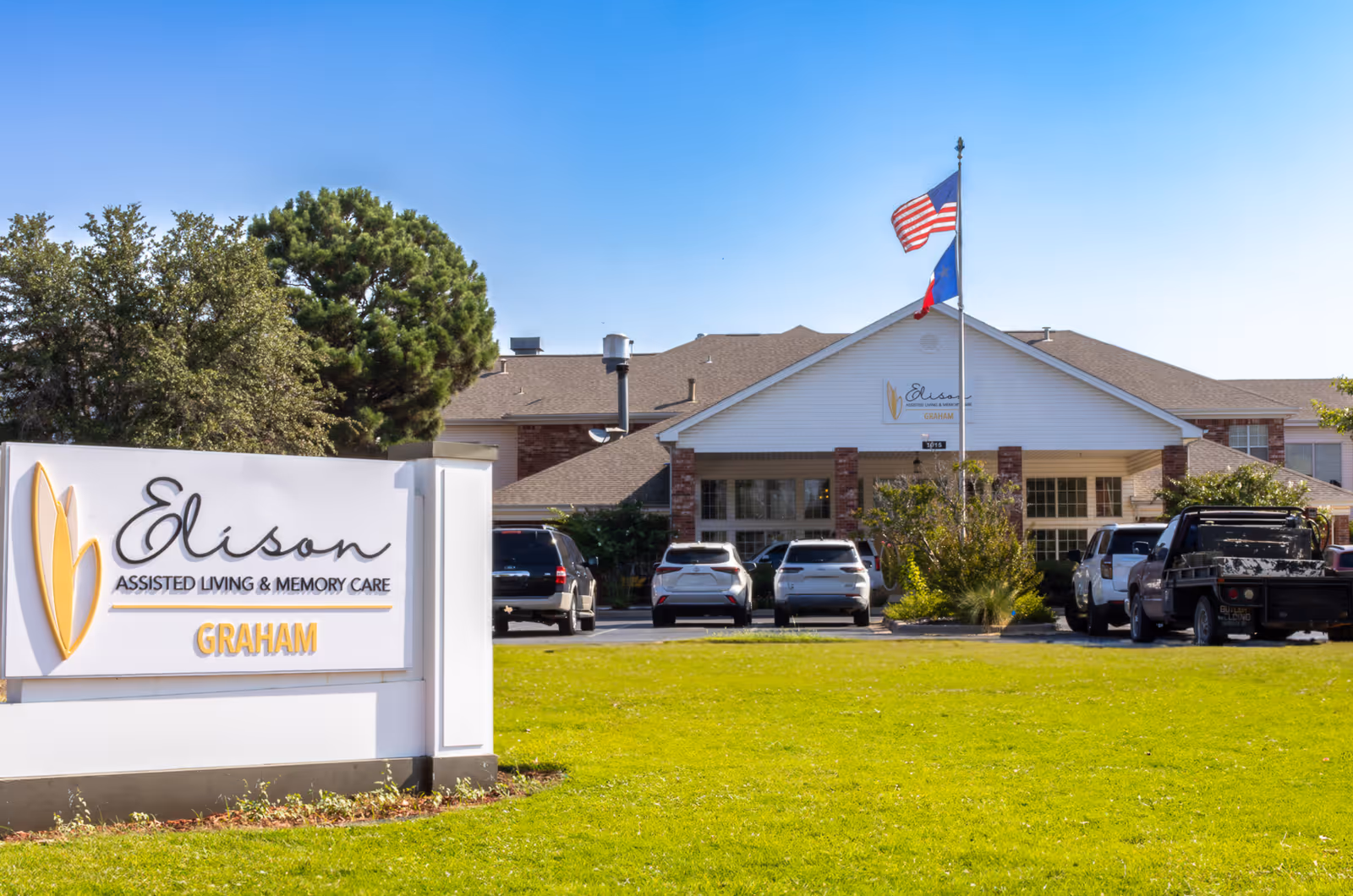 Front exterior of Elison Assisted Living & Memory Care of Graham showing the entrance, facility sign, parked cars, and flagpoles on a grassy lawn.