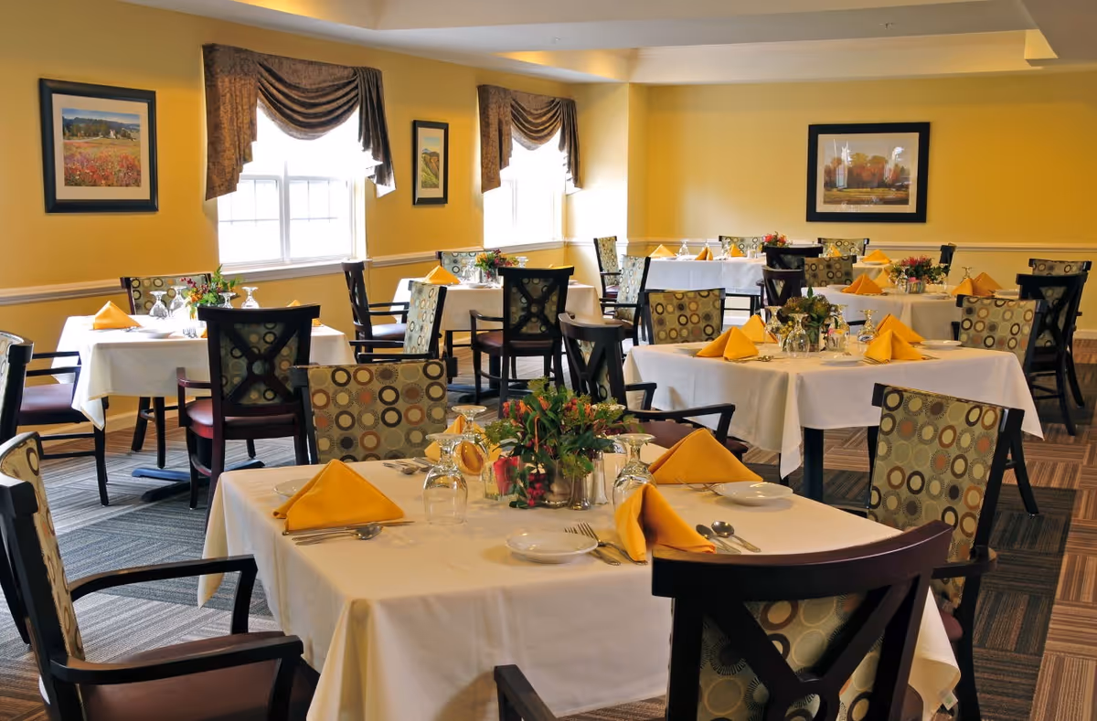 A dining room with multiple tables covered in white tablecloths, each set with yellow folded napkins, plates, glasses, and silverware. The chairs have patterned upholstery with circular designs. The walls are painted yellow and decorated with framed artwork. Two windows with brown curtains allow natural light into the room.