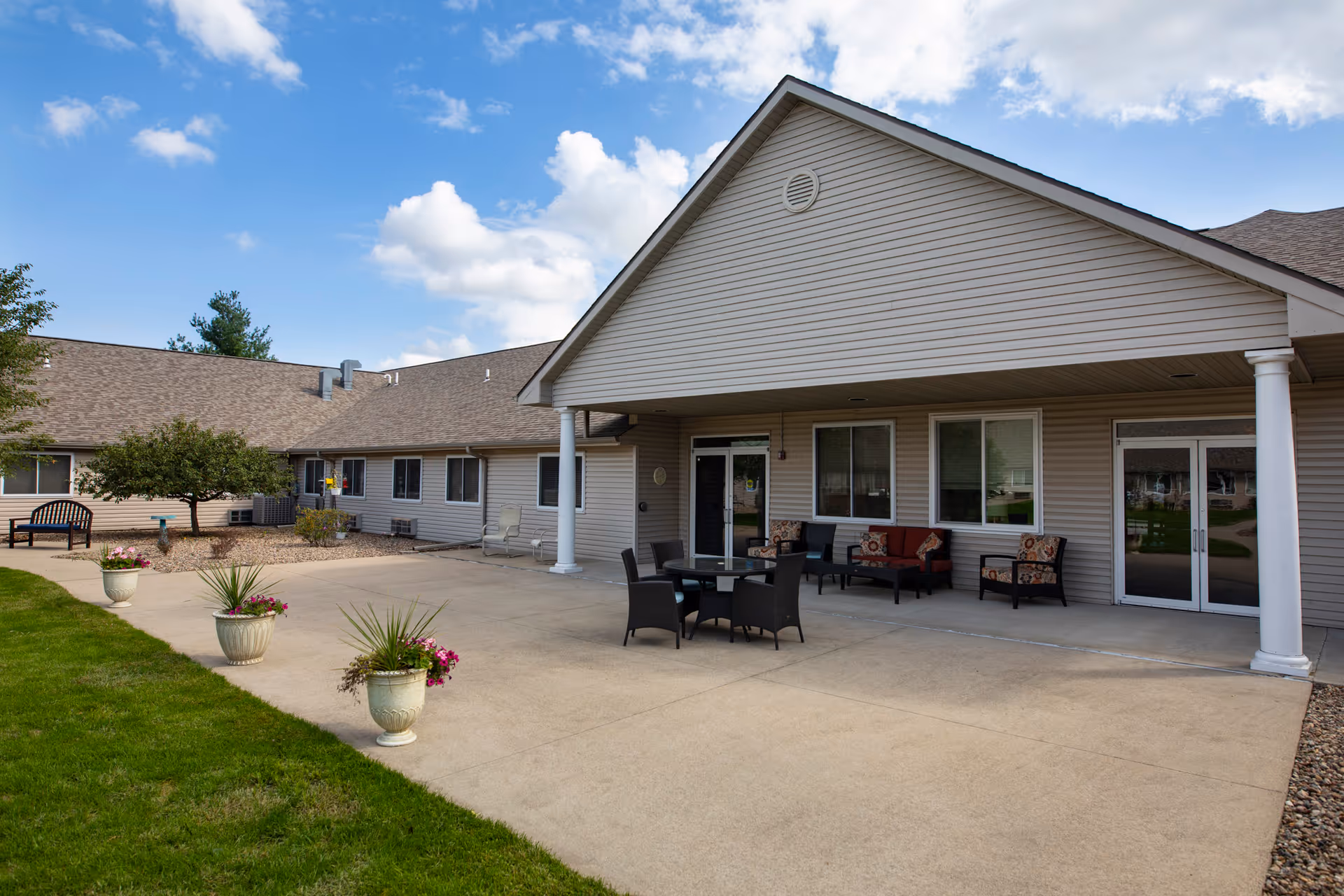 Outdoor patio area of a single-story assisted living facility with beige siding and a gabled roof. The patio features a round table with four chairs, cushioned seating along the wall, potted plants, and a bench near a small tree. The sky is partly cloudy.