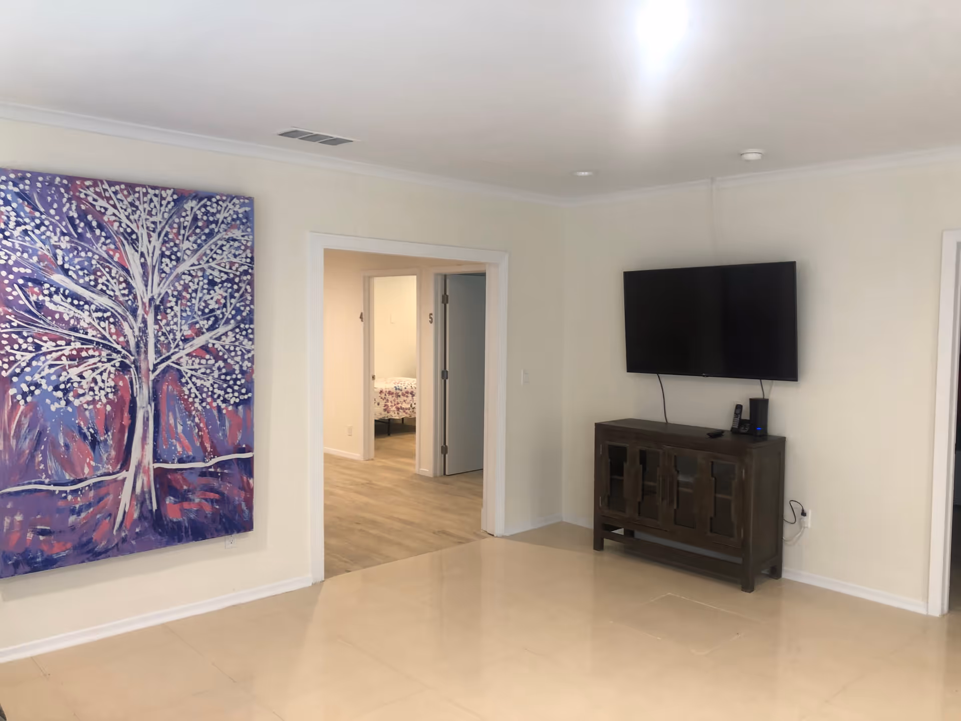 A clean, minimally furnished room with cream-colored walls and tiled floor. A large colorful painting of a tree with white blossoms on a purple and pink background hangs on the left wall. A flat-screen TV is mounted on the right wall above a dark wooden cabinet. An open doorway in the center leads to a hallway with light wood flooring and visible doors, one of which reveals a bed with floral bedding.
