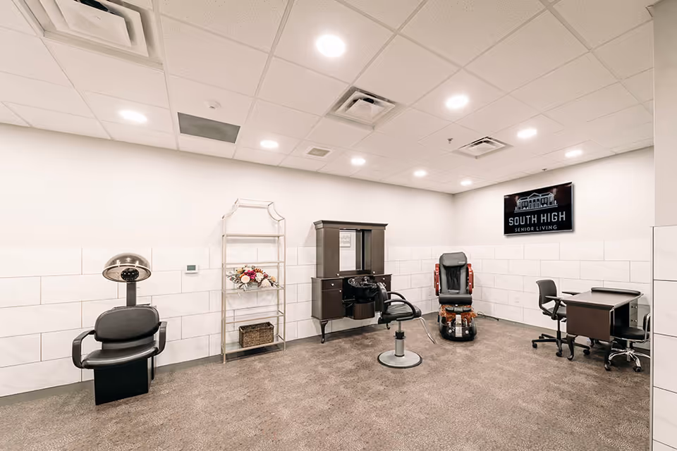 A spacious salon room in South High Senior Living with a hair washing station, a styling chair in front of a mirror cabinet, a pedicure chair, a desk with two office chairs, and a metal rack holding flowers and a basket. The walls are white with tile halfway up, and the ceiling has recessed lighting. A sign on the wall reads 'South High Senior Living.'
