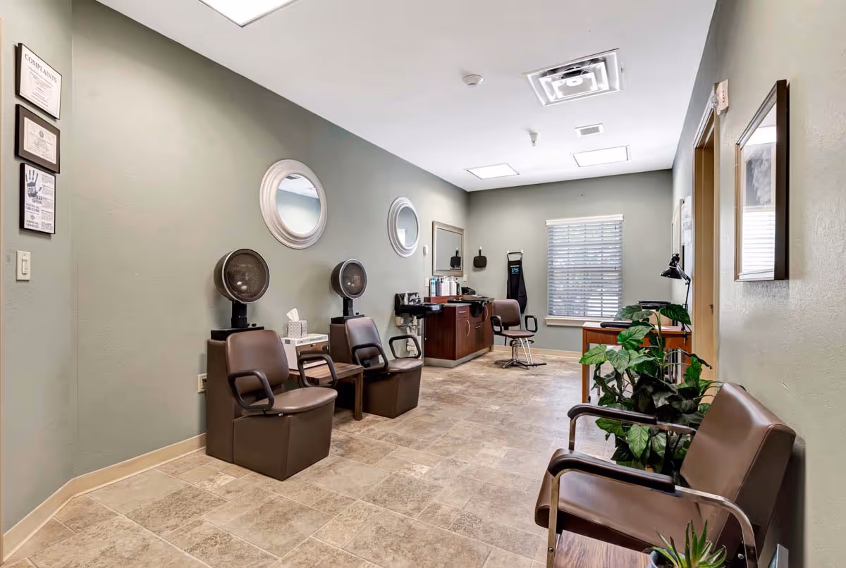 Interior view of a salon area in a senior living facility with two brown salon chairs under hair dryers, a third salon chair near a mirror and counter with hair products, a window with blinds, a couple of framed notices on the wall, and a couple of brown chairs with plants nearby.