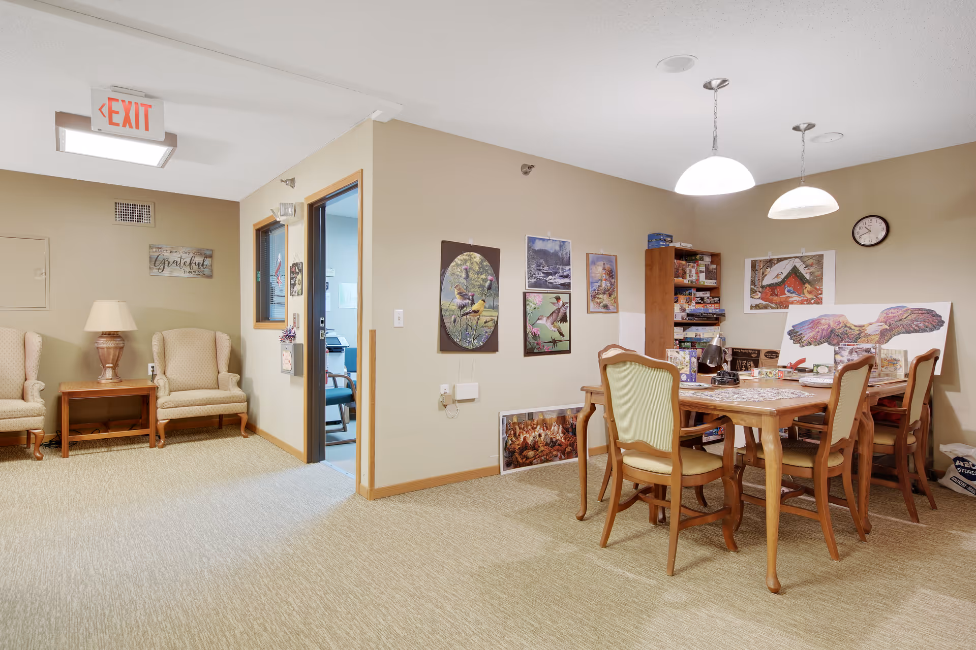 A cozy common area in Elmhurst Commons featuring a wooden table with six chairs, two hanging pendant lights, and various framed artwork and puzzles on the walls and table. To the left, there are two beige armchairs with a wooden side table and lamp between them, under an exit sign and a wall decoration that says 'Grateful'. The room has beige walls and carpeted flooring.