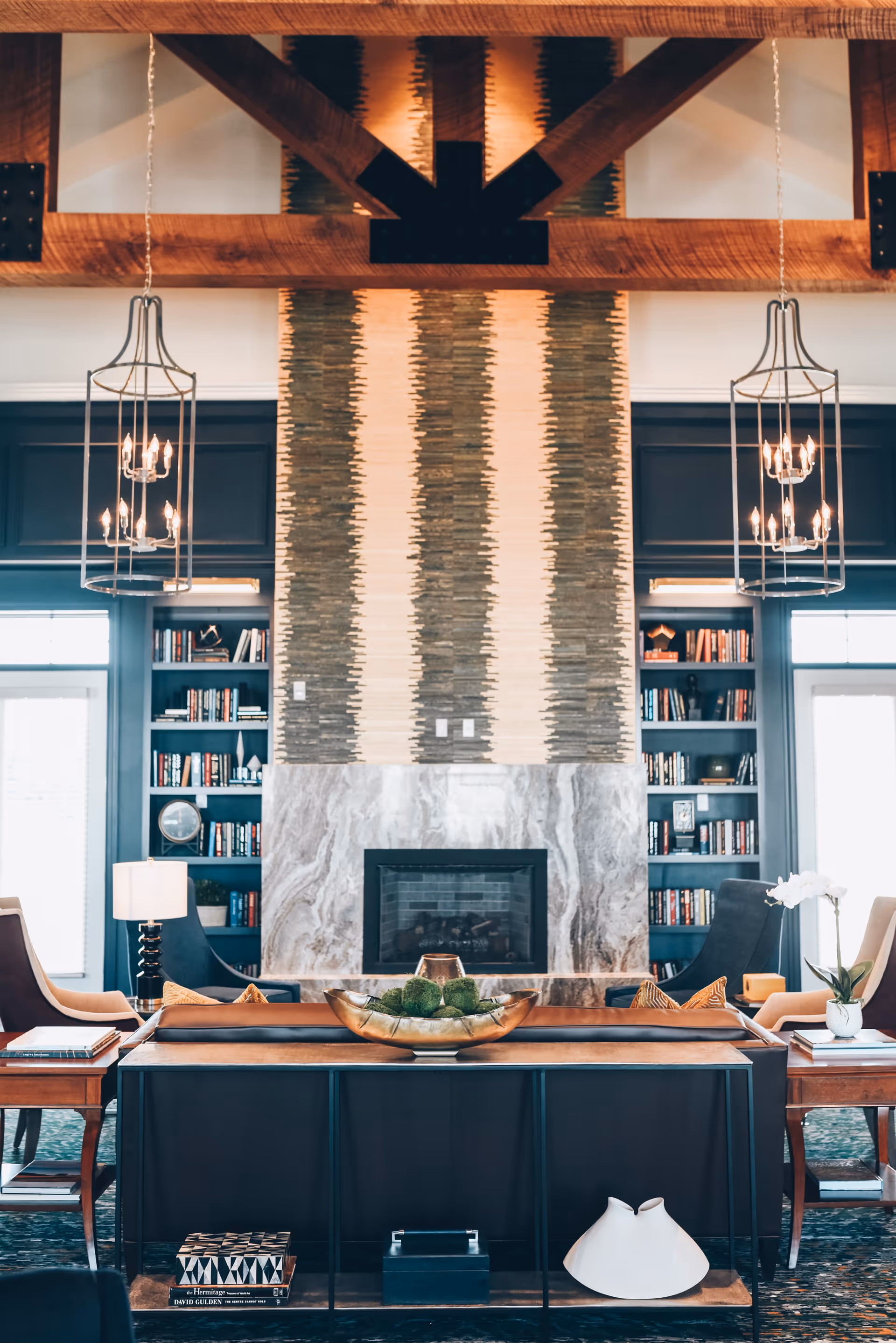 High-ceilinged lounge with a central fireplace flanked by built-in bookshelves, seating, chandeliers, and decorative accents.