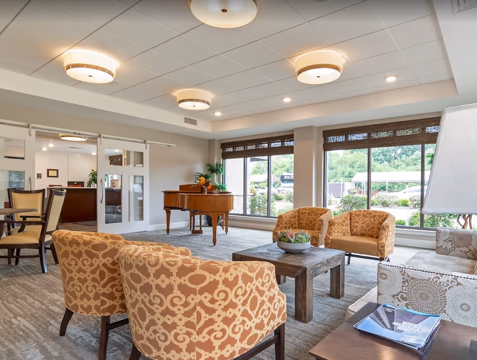 A bright and spacious senior living facility common area with large windows letting in natural light. The room features patterned armchairs arranged around a wooden coffee table with a small plant centerpiece. A grand piano is positioned near the windows, and there are additional chairs and tables in the background. The ceiling has modern round light fixtures, and the overall decor is warm and inviting.