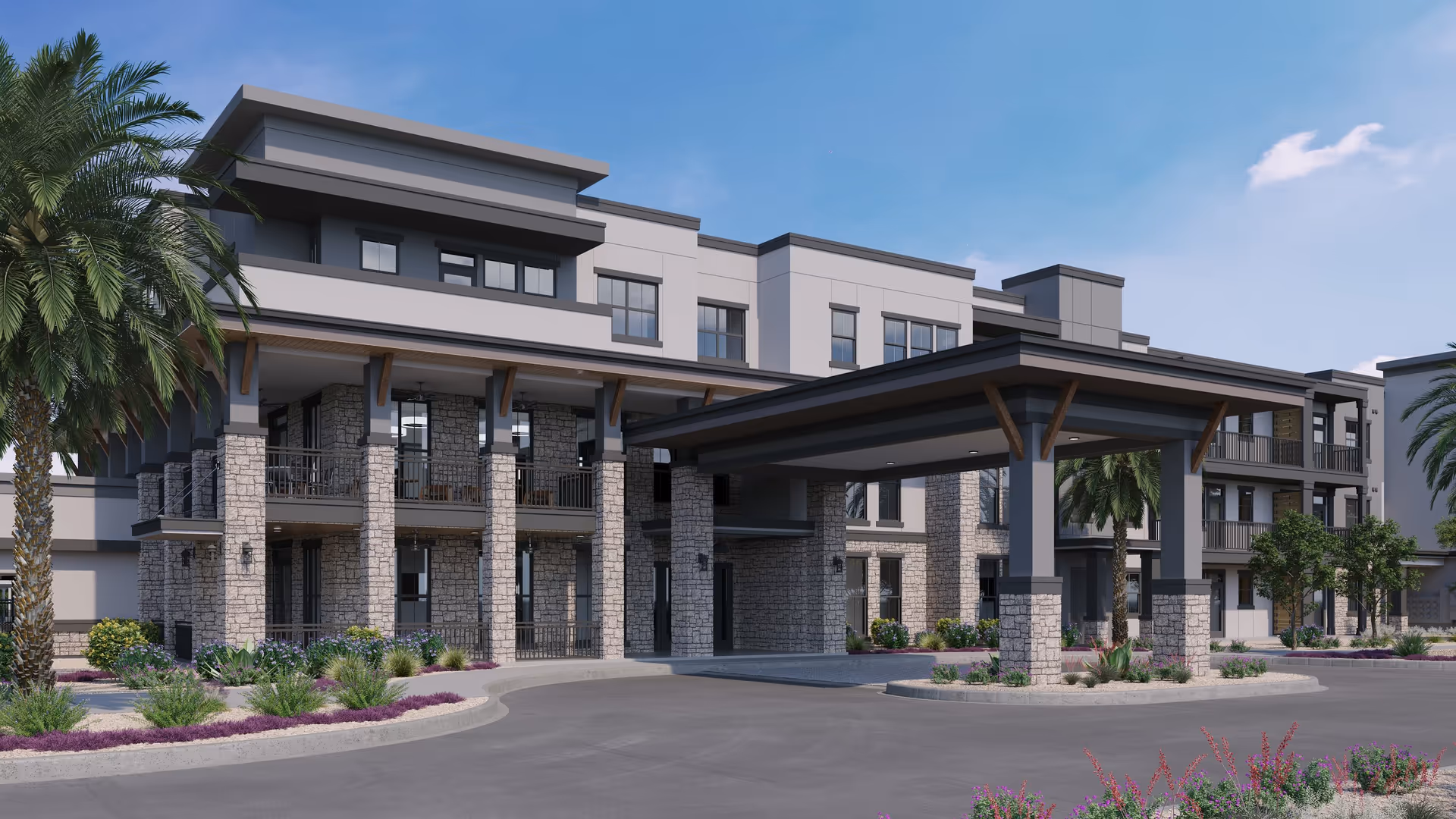 Exterior view of a modern senior living facility building with stone pillars, balconies, and a covered entrance driveway. The landscaping includes palm trees, shrubs, and flowering plants under a clear blue sky.