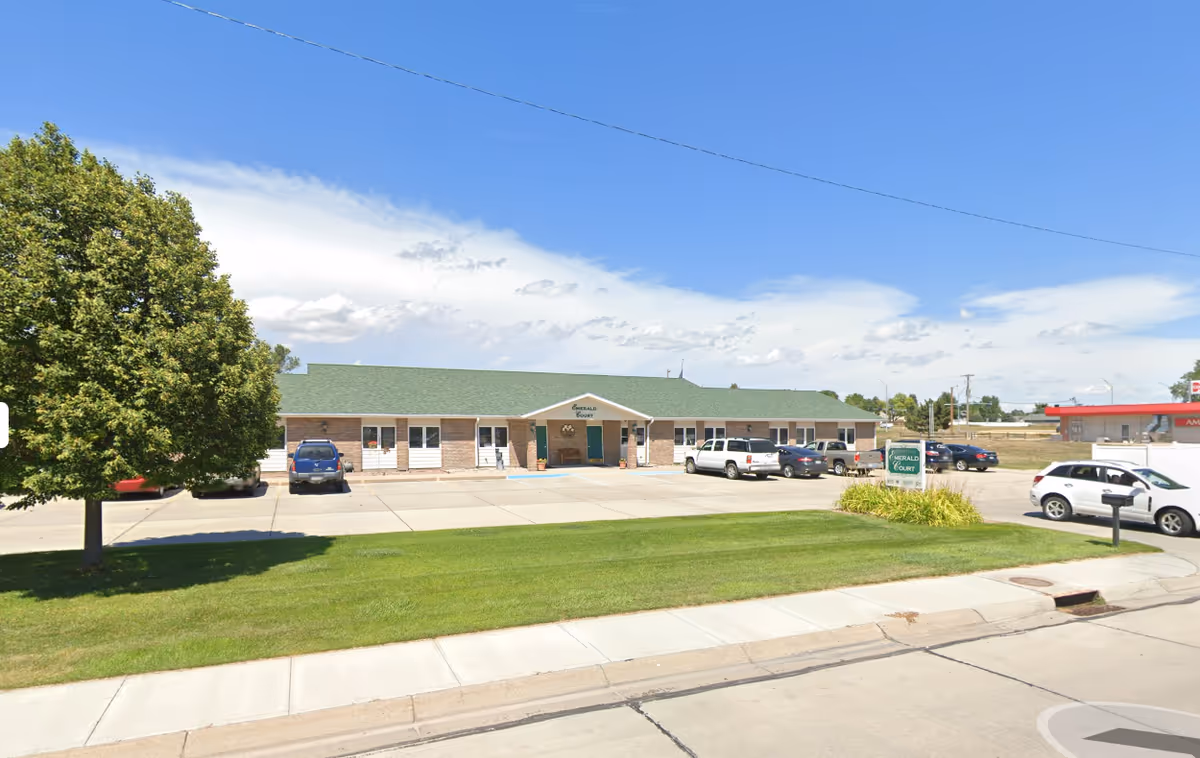 Exterior view of Emerald Court, a single-story brick building with a green roof, several parked cars in front, a green lawn, and a tree on the left side under a partly cloudy blue sky.