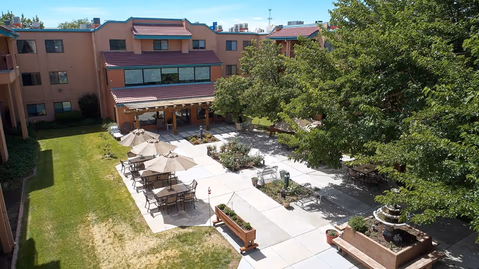 Outdoor courtyard area of Sandia Vista Senior Living featuring patio tables with umbrellas, benches, a water fountain, and surrounding greenery with trees and grass, adjacent to a multi-story building with large windows and a covered entrance.