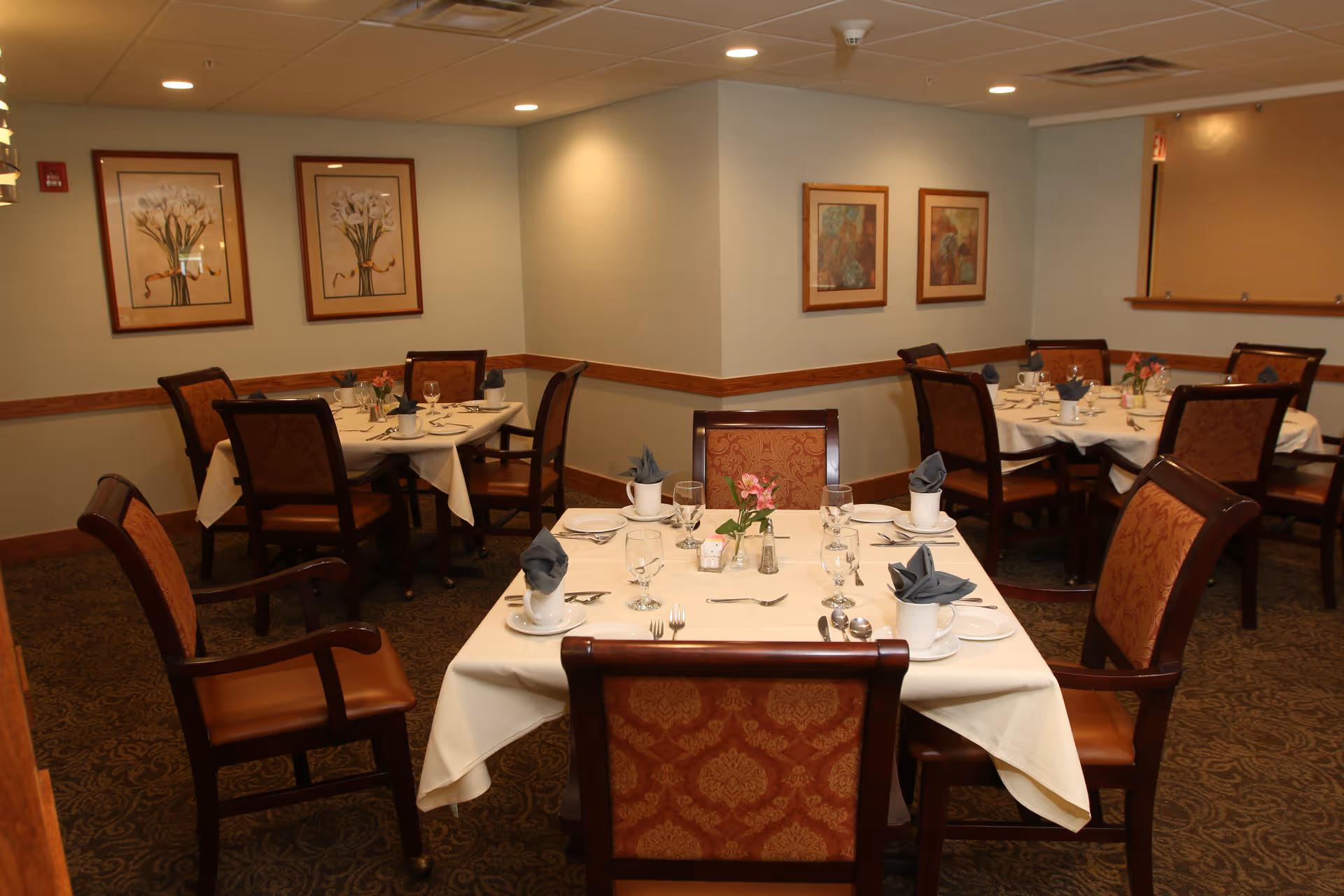 A dining room with several tables set for a meal. Each table is covered with a white tablecloth and set with plates, glasses, silverware, and napkins folded in cups. The chairs have wooden frames with patterned upholstery. The walls are decorated with framed floral artwork, and the room has soft lighting.