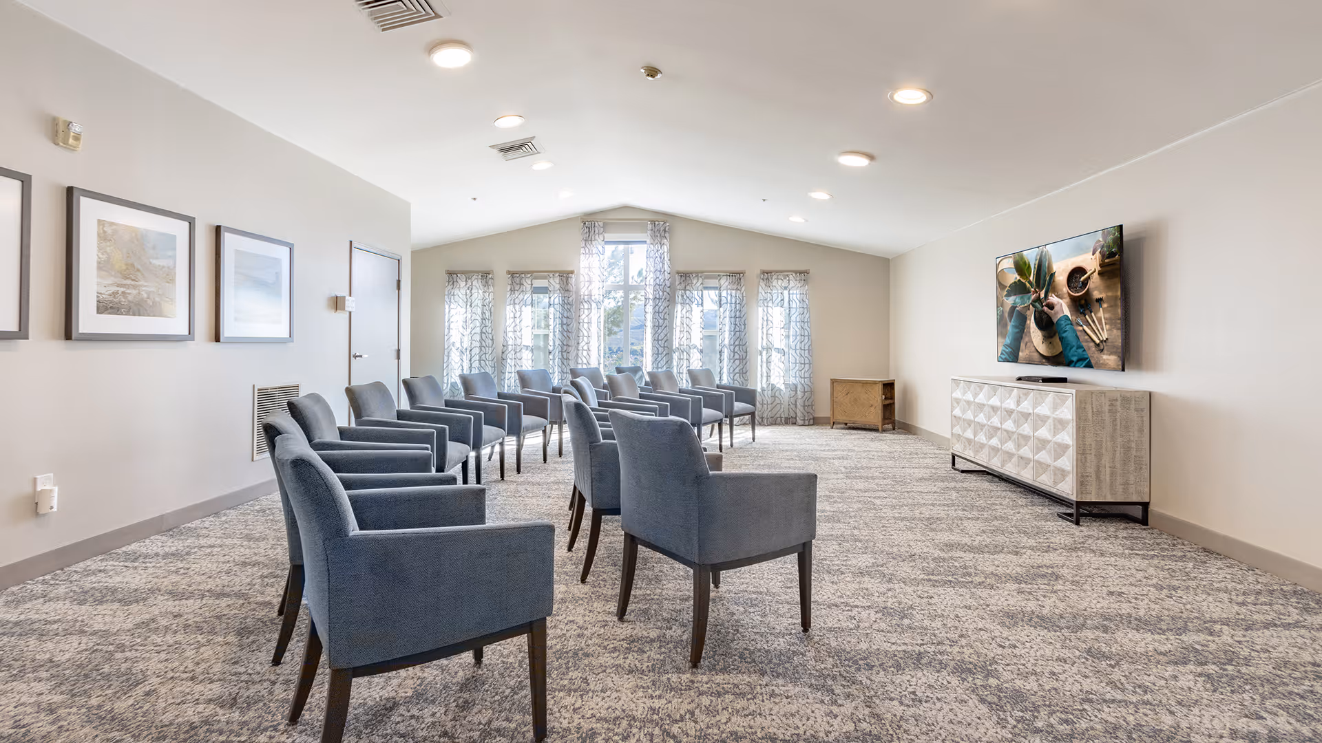 Bright communal lounge with rows of blue upholstered chairs facing a wall-mounted TV and windows.