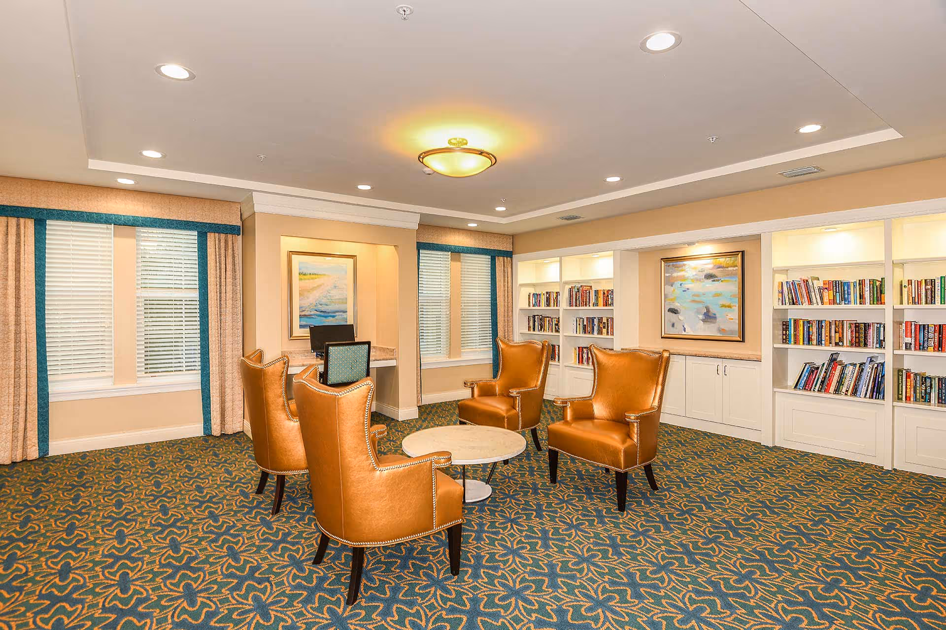 A cozy sitting area in a senior living facility with four brown leather armchairs arranged around a round white table. The room features patterned carpet, built-in white bookshelves filled with books, two framed paintings on the walls, and windows with beige and teal curtains. There is also a small desk with a computer in a recessed wall area.