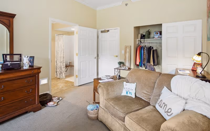 A cozy living area with a brown sofa adorned with decorative pillows, a wooden dresser with a mirror, and an open closet with hanging clothes. The room has beige walls and carpeted floor. There is a doorway leading to a bathroom with a shower curtain visible.