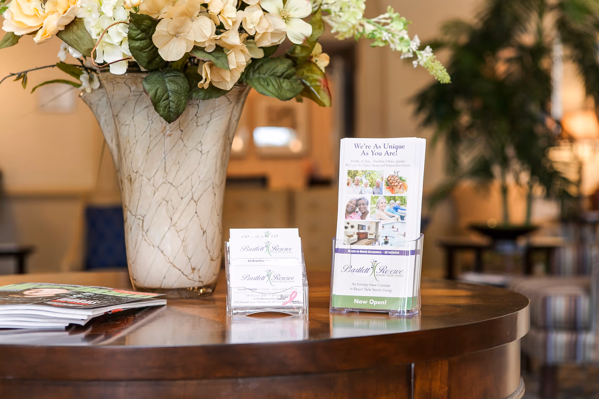 A close-up view of a wooden table with a large decorative vase filled with white and green flowers. On the table are brochures and business cards for Bartlett Reserve, a senior living facility. The background shows a softly lit, cozy interior with plants and furniture.