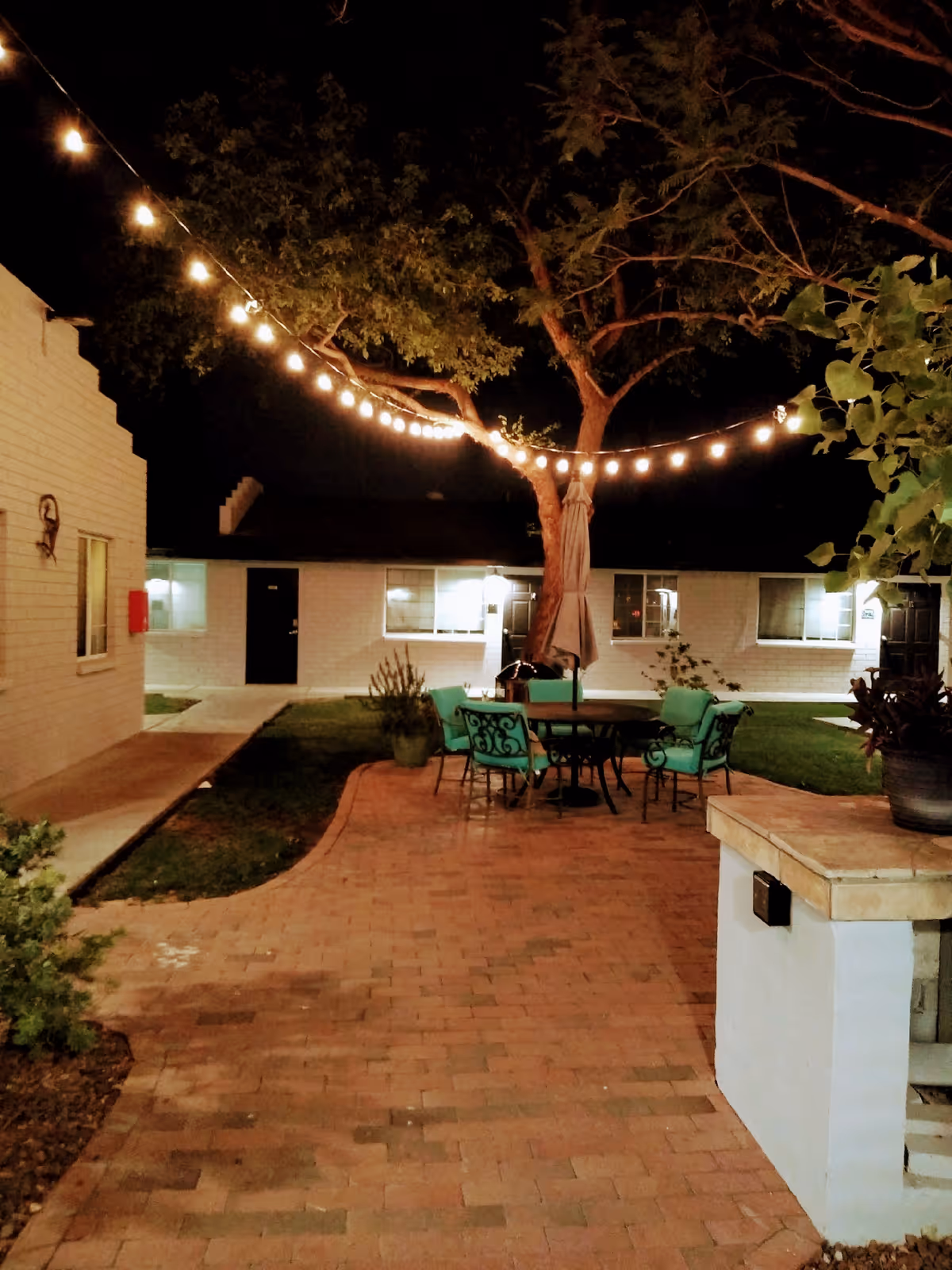 Nighttime courtyard with string lights, a table and turquoise chairs under a tree on a brick-paved area surrounded by single-story buildings.