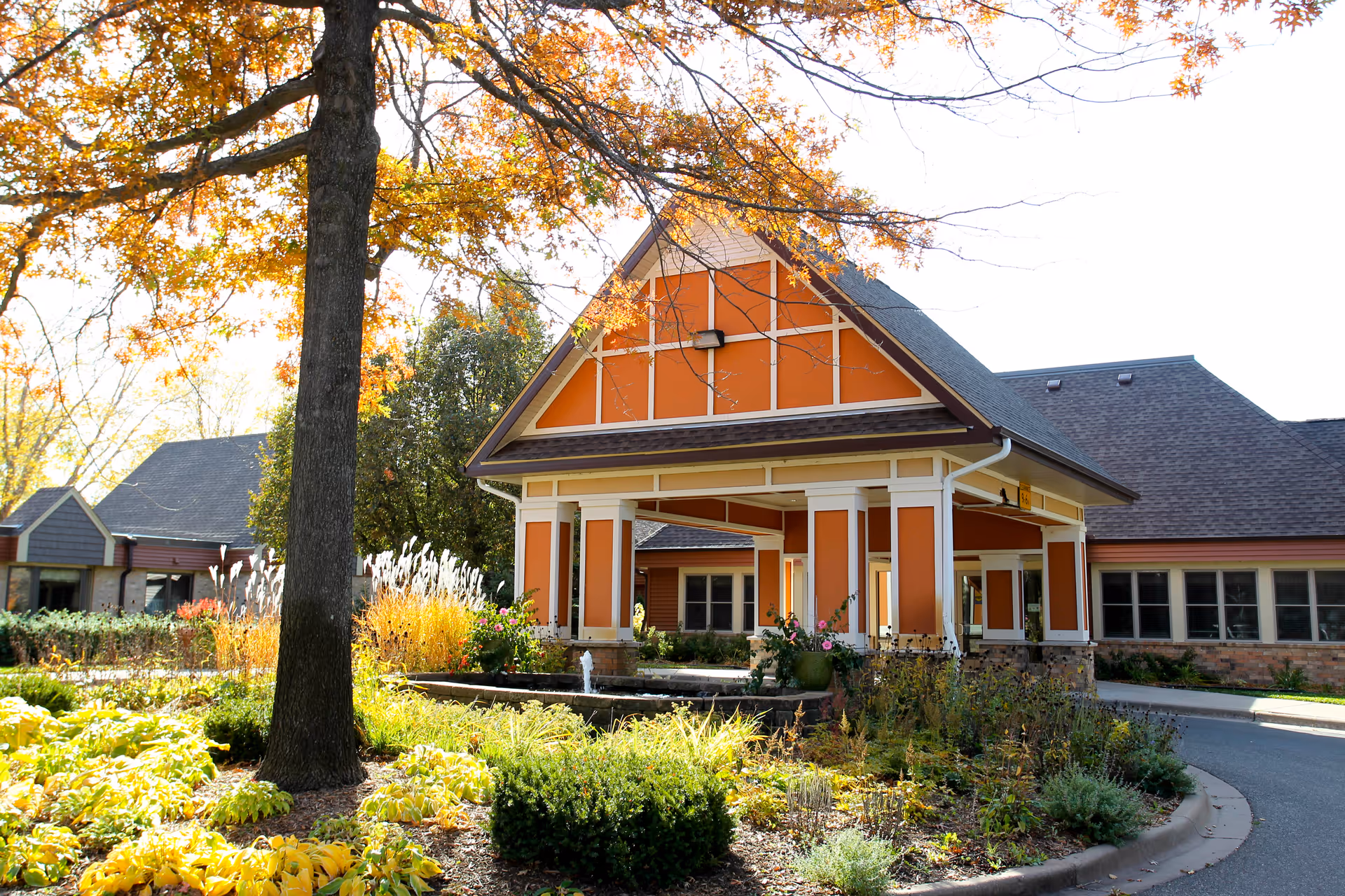 Exterior view of Ecumen Point Pleasant Heights facility showing a covered entrance with orange and white trim, surrounded by landscaped gardens and trees with autumn foliage.