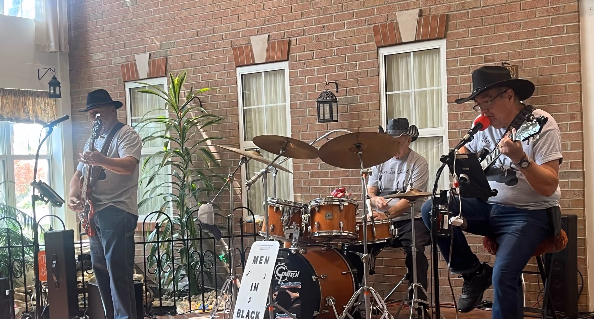 A three-member band performing indoors in front of a brick wall with windows. The band members are playing guitar and drums, wearing casual clothes and black hats. There is a sign in front of the drum set that reads 'MEN IN BLACK'. The setting appears to be a common area with plants and natural light coming through a window on the left.