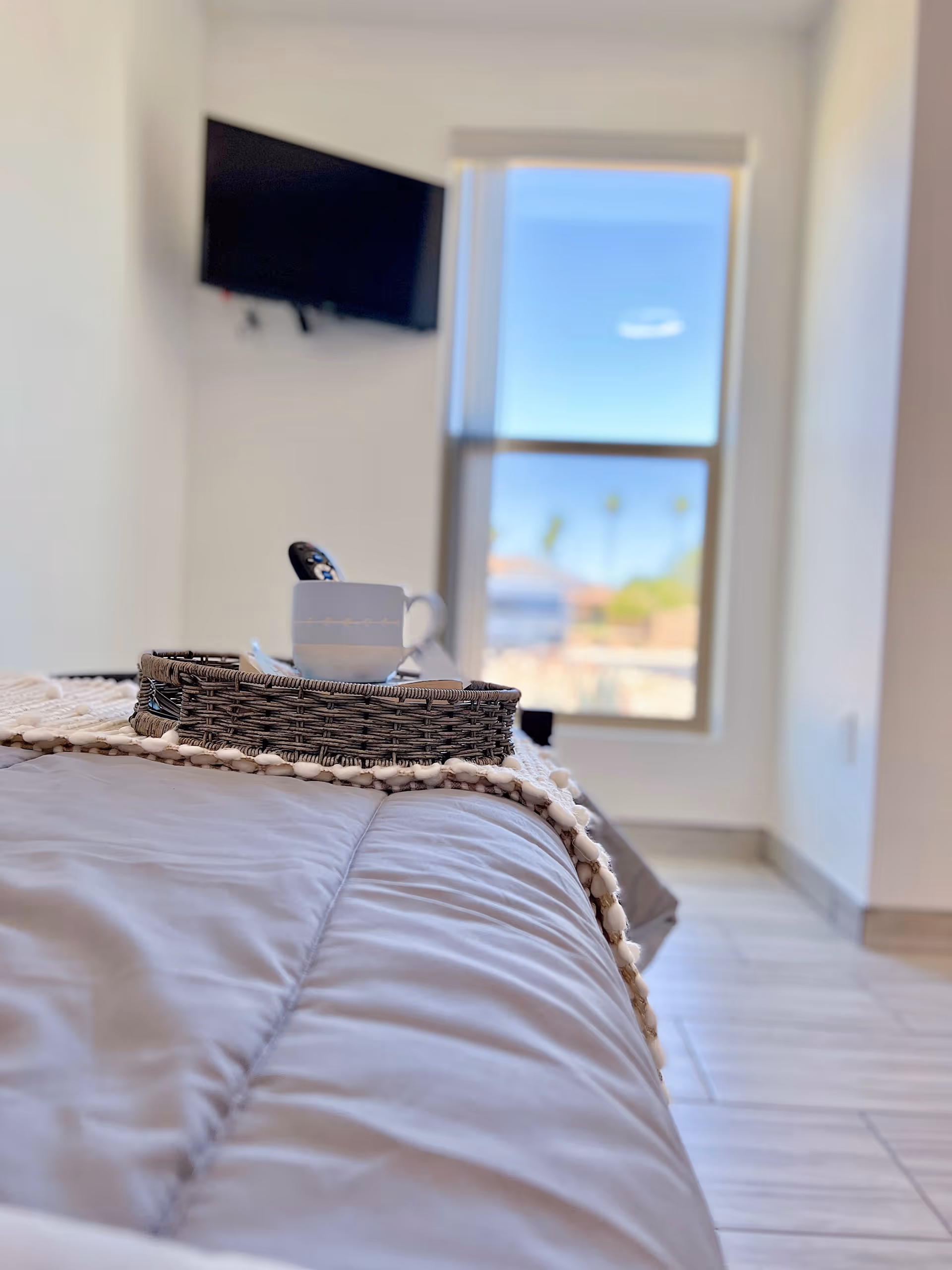 Close-up view of a bed with a gray comforter and a decorative throw blanket. On the bed is a woven tray holding a white cup and a remote control. In the background, there is a window showing a clear blue sky and some palm trees, and a wall-mounted TV is visible on the left side.
