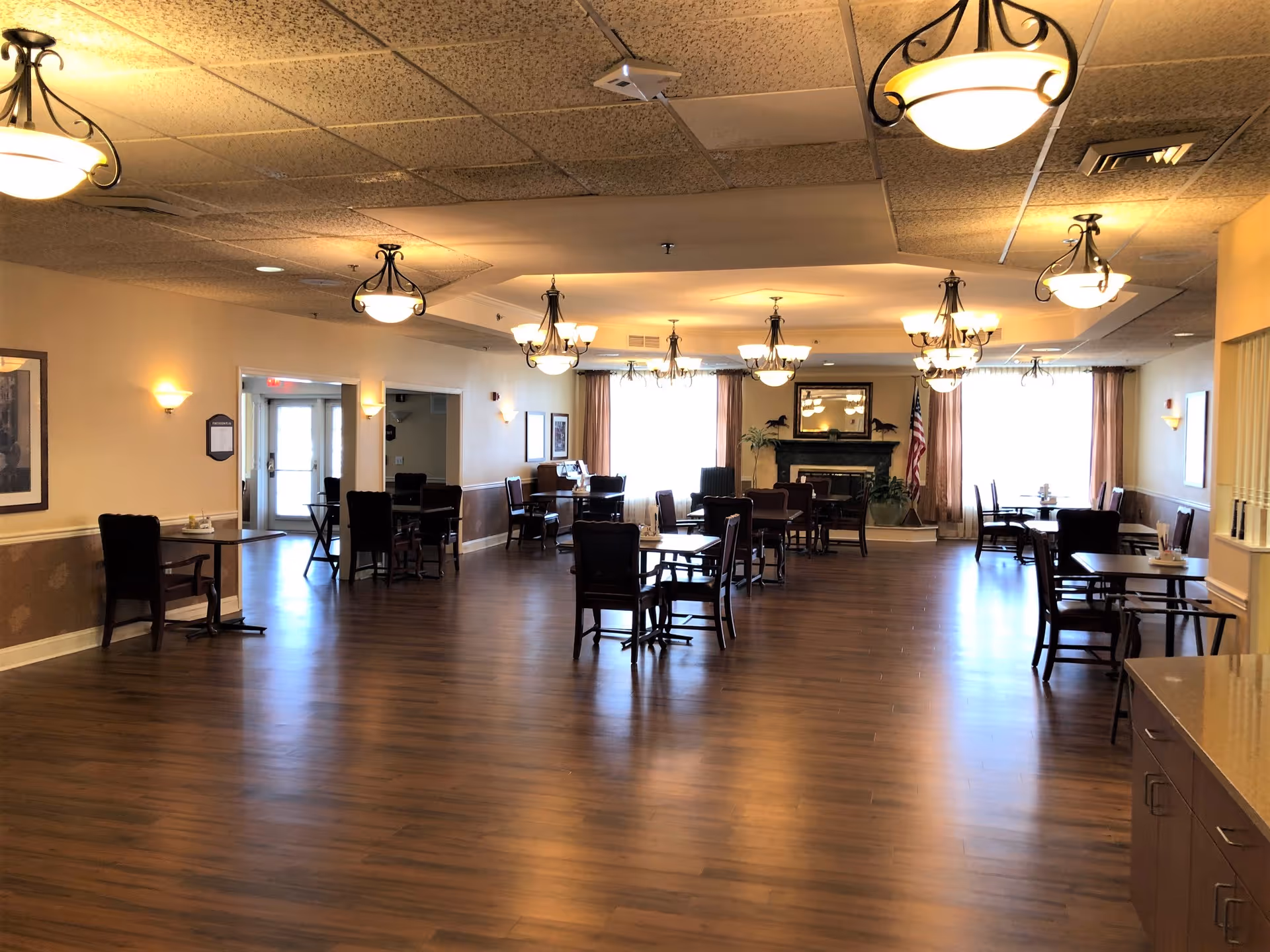Spacious dining/activity room with wooden floors, multiple tables and chairs, chandeliers, and a fireplace at the far end.