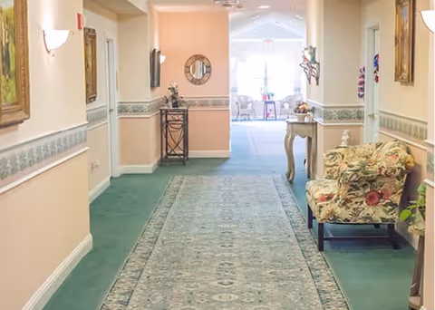 A well-lit hallway in an assisted living facility with green carpet and a patterned runner rug. The walls are painted peach with a decorative border and adorned with framed artwork. There is a floral upholstered armchair and a small table with a lamp on the right side. At the end of the hallway, there is a sitting area with chairs and a table near large windows letting in natural light.