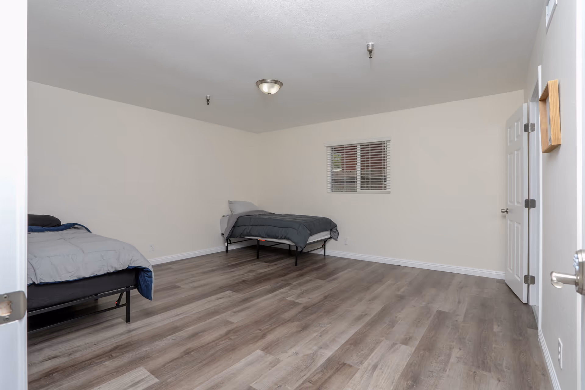 A simple bedroom with two single beds, each with gray bedding, positioned against cream-colored walls. The room has a window with white blinds, wood-look flooring, and a ceiling light fixture. A white door is open on the right side of the room.