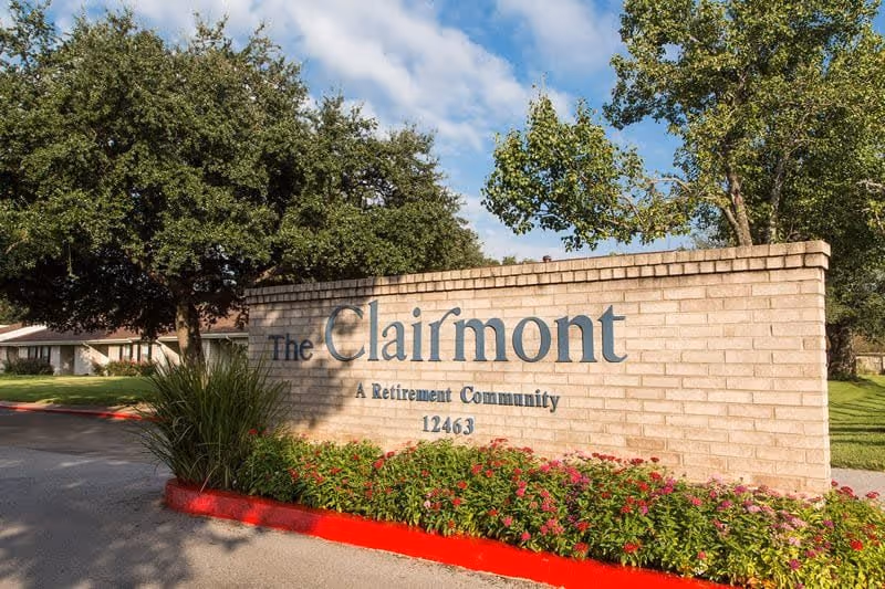 Brick entrance sign reading "The Clairmont A Retirement Community 12463" with landscaping, trees, and a blue sky.