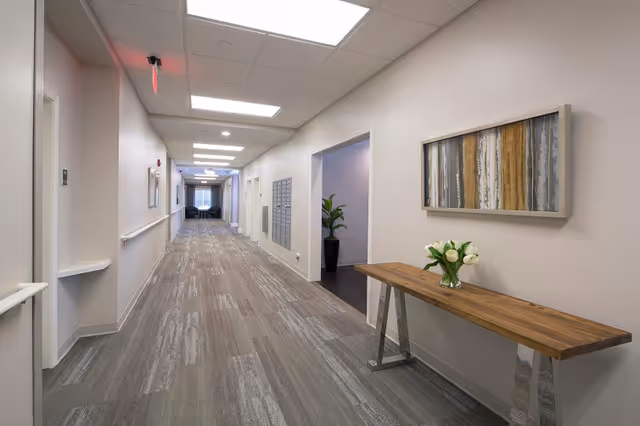 A long, well-lit hallway in a senior living facility with light-colored walls and patterned carpet. On the right side, there is a wooden console table with a vase of white tulips and a framed abstract artwork above it. The hallway has several doors and a seating area visible at the far end.