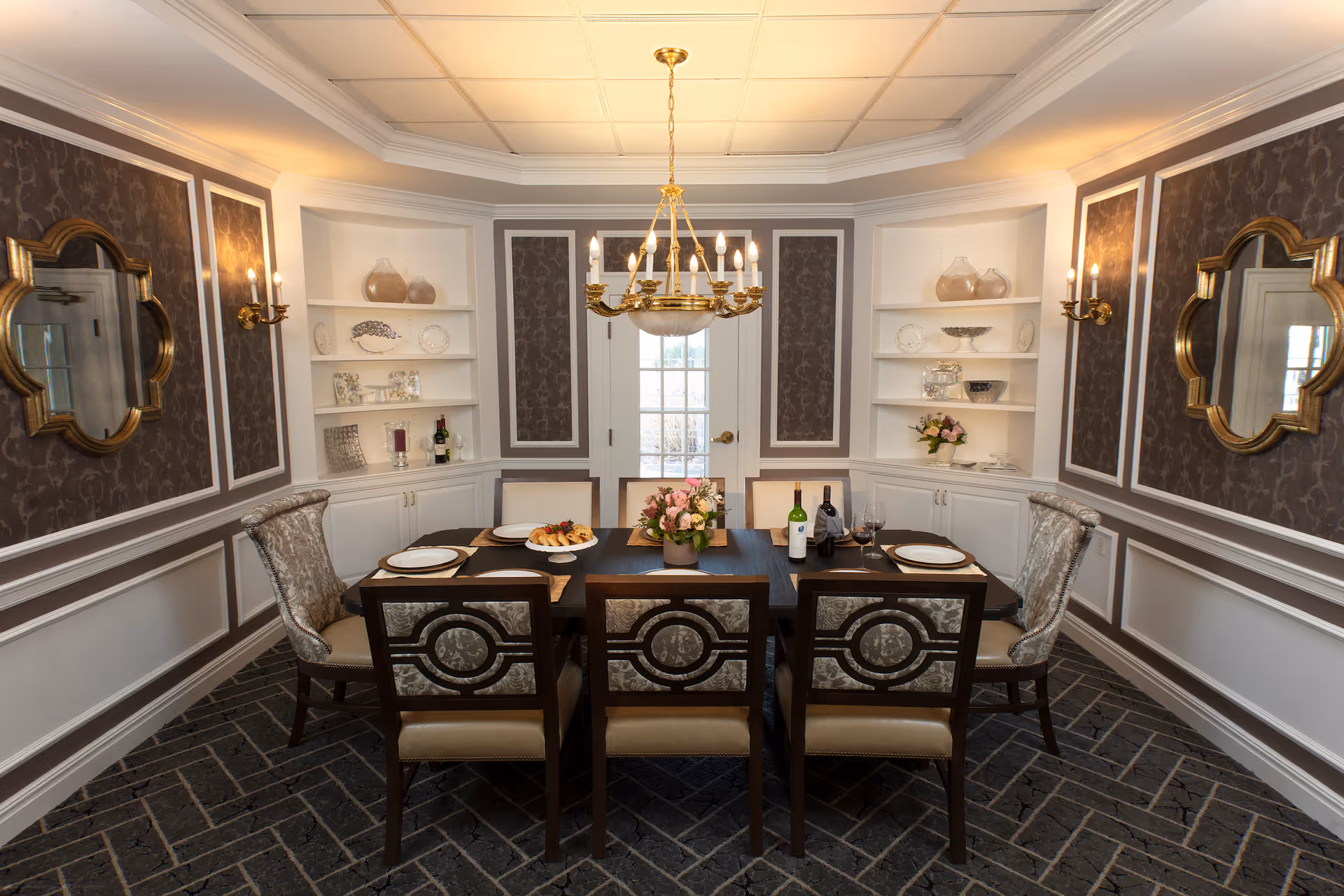 Elegant dining room with a dark wooden table set for six, featuring patterned upholstered chairs. The room has decorative wallpaper panels, two gold-framed mirrors on opposite walls, built-in white shelves with decorative items, and a gold chandelier hanging from a tray ceiling. The floor is covered with dark patterned carpet.