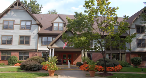 Exterior view of a multi-story senior living facility building with a brick and siding facade, multiple windows, a central entrance with a small porch, an American flag, and landscaped greenery including trees, bushes, and potted plants.
